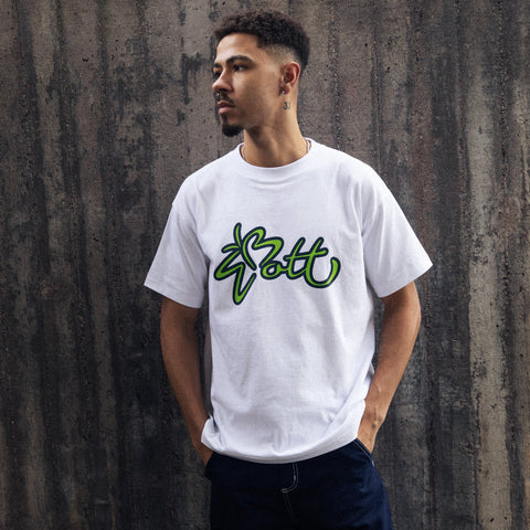 Man wearing a white t-shirt with a large green and black script logo reading "Bott", standing against a dark concrete wall with hands in pockets.