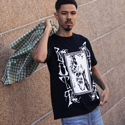 Man wearing a black graphic t-shirt with large white gothic lettering and a framed white skull-like print, a plaid shirt slung over one shoulder, against a tan stone wall.