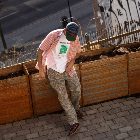Person leaning against a rusted metal planter wearing a pink gingham shirt, white graphic tee, camouflage cargo pants, brown shoes, and a patterned cap.