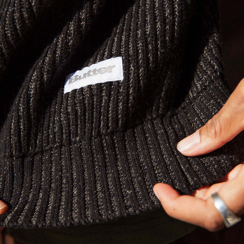 Close-up of a black ribbed knit sweater with a white woven label reading "Butter" and a hand with a silver ring at the hem.