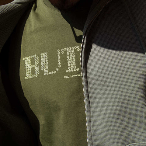 Close-up of a person wearing an olive green crew-neck tee with a dotted white graphic logo and a URL, layered under a grey waffle-knit zip jacket.