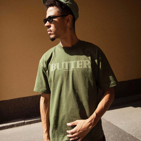 Man in sunglasses and a green cap wearing an olive green t-shirt with the word BUTTER in light lettering, standing against a tan wall.