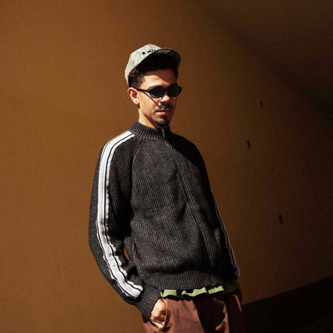 Man in dark knit sweater with white side stripes, sage green cap, and sunglasses, standing against a warm brown wall.