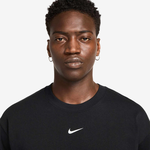Man wearing a black crew-neck t-shirt with a small white Swoosh logo on the chest, and small hoop earrings, against a plain white background.