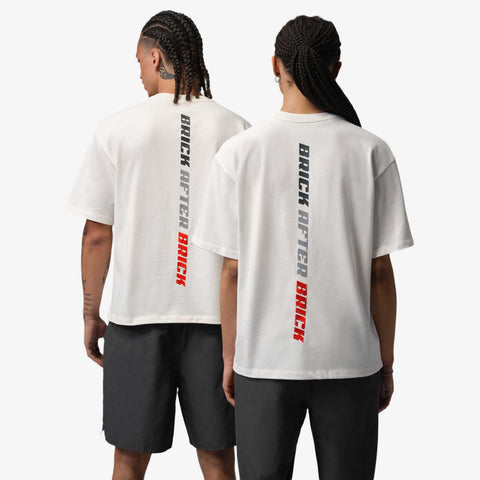 Two people seen from behind wearing white oversized t-shirts with vertical 'BRICK AFTER BRICK' text in black and red on the back, paired with dark grey shorts.