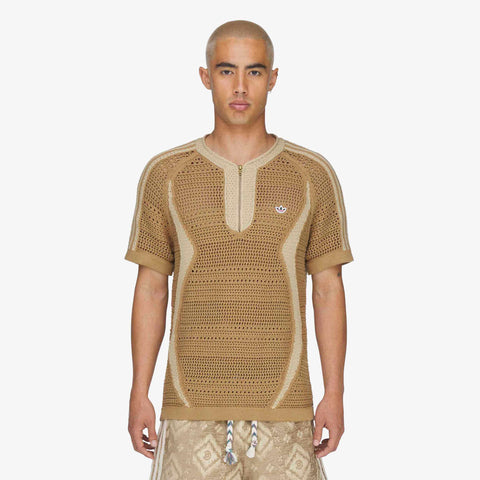 Model wearing a tan open-knit short-sleeve henley top with a zip placket, contrast beige trim, and a small embroidered logo on the chest, paired with patterned shorts.