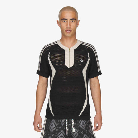 Model wearing a black open-knit short-sleeve top with cream trim, a half zip collar, and a small logo on the chest, with patterned shorts.