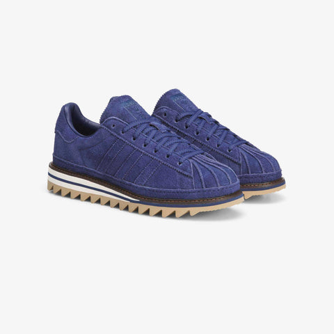 Pair of navy blue suede low-top sneakers with a shell-toe cap, white stripes and a serrated gum-and-white rubber sole, shown from a front three-quarter angle.