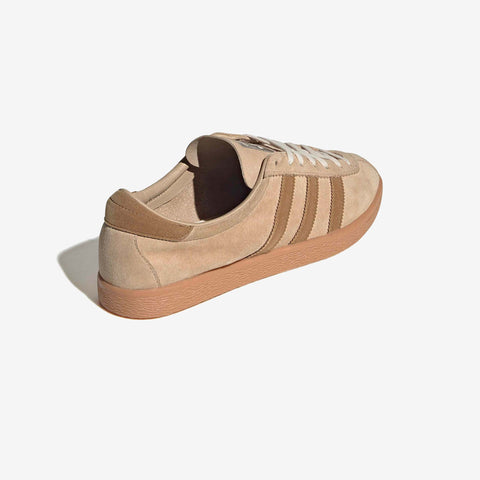 Tan suede low-top sneaker with brown three-stripe side panels, cream laces, and a gum rubber sole, shown from the rear three-quarter angle.