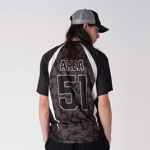 Person seen from behind wearing a black short-sleeve jersey with white raglan panels, a brown skull pattern, and 'AREA 51' printed on the back, plus a grey and black cap.