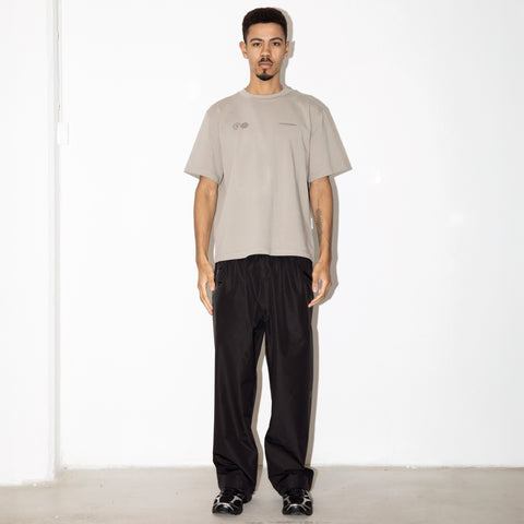 Man standing against a white wall wearing a light grey short-sleeve t-shirt and black wide-leg pants with black shoes.