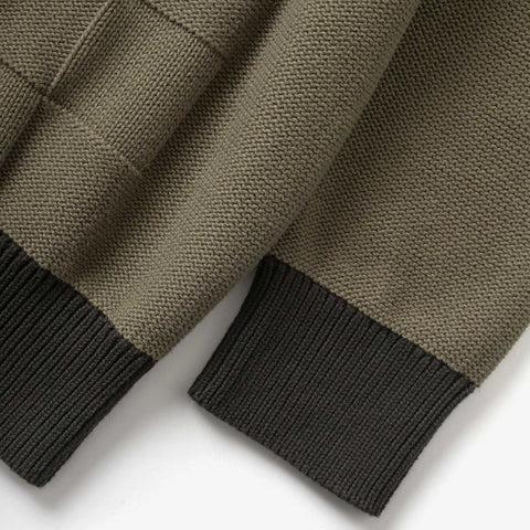 Close-up of two olive green knit sweater sleeves on a white surface, each ending in a dark charcoal ribbed cuff.