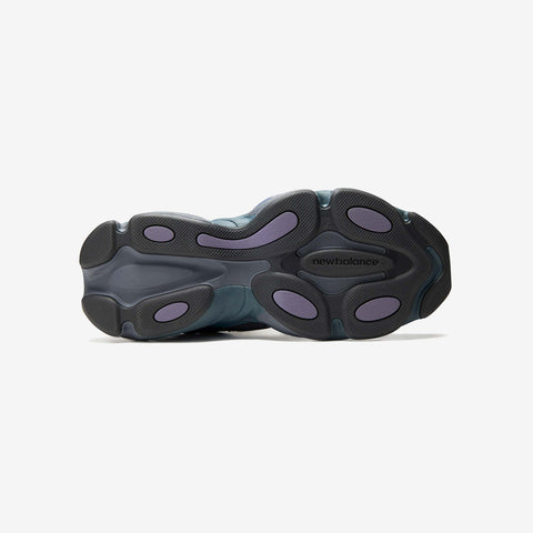 Underside of a grey and purple New Balance running shoe, showing a sculpted black rubber outsole with purple pods and the brand name printed on the sole.
