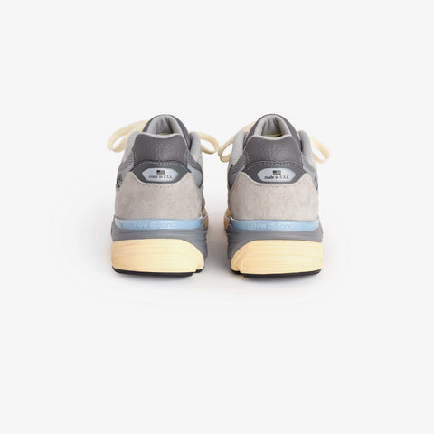 Rear view of a grey suede running shoe with cream laces, light blue accents, and a cream-colored midsole on a white background.