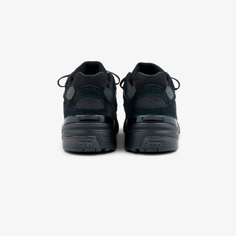 Rear view of a pair of black suede low-profile running sneakers with black laces and a thick black rubber sole, on a white background.