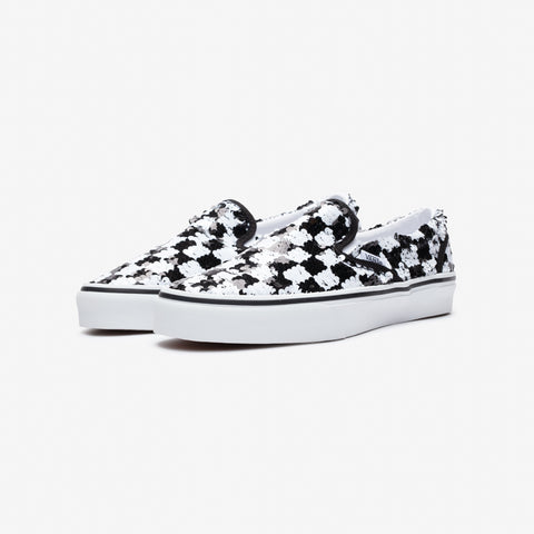Pair of black and white slip-on sneakers with a checkered pattern of splattered paint and a white rubber sole, shown from the front three-quarter angle.