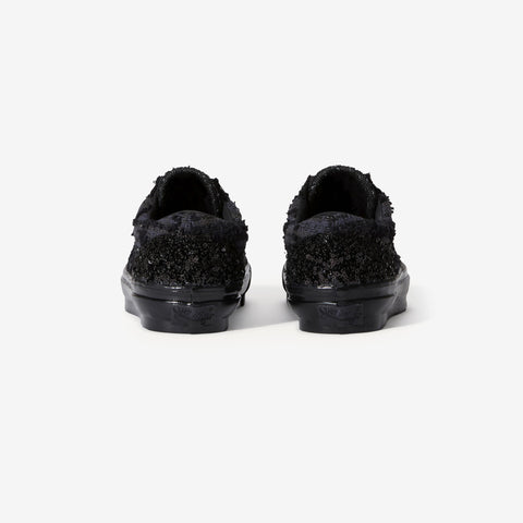 Rear view of a pair of black low-top sneakers with a textured, shaggy black upper and black rubber soles on a white background.