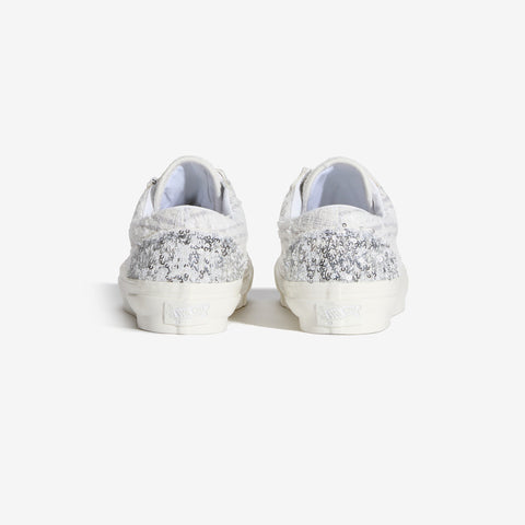 Rear view of a pair of white low-top sneakers with a silver sequined and lace-textured upper and white rubber soles.