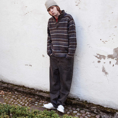 Person leaning against a white wall wearing a grey knit beanie, a striped half-zip pullover in brown, grey and blue, dark wide-leg trousers and white sneakers.