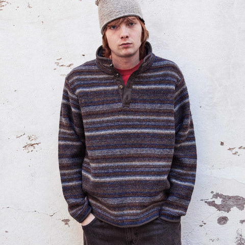 Young man in a grey knit beanie and a striped brown, blue and grey henley-neck sweater with a red shirt underneath, hands in pockets against a white wall.