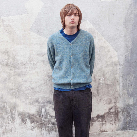 Person standing against a weathered concrete wall, wearing a light blue knit cardigan over a blue crew-neck top and dark grey wide-leg trousers.