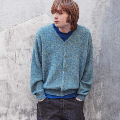 Young man with shoulder-length hair wearing a fuzzy teal-blue knit cardigan over a blue shirt, hands in pockets, against a grey concrete wall.